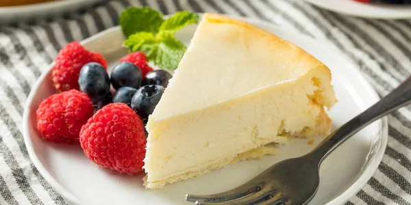 A plated cheesecake slice with graham cracker crust, fresh raspberries, blueberries, and mint garnish for foodservice and wholesale bakery supply.