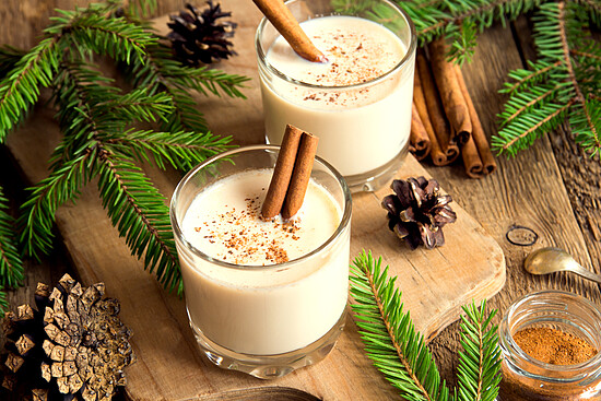 Creamy eggnog with cinnamon sticks, surrounded by evergreen branches and rustic pine cones.