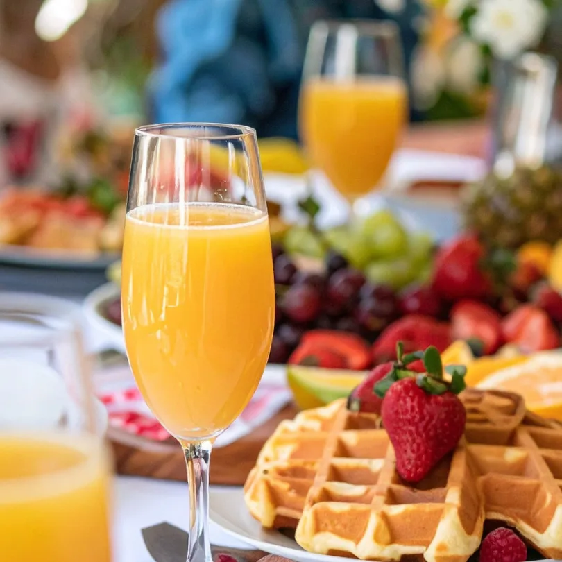 Elegant champagne flute filled with fresh orange juice surrounded by gourmet breakfast items including waffles, fresh berries, and seasonal produce for foodservice wholesale.