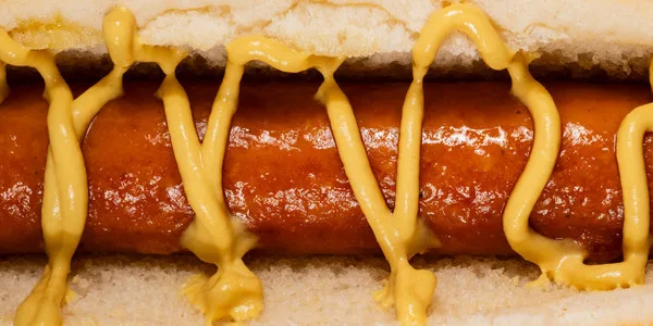 Professional food service hot dog on toasted bun with decorative yellow mustard stripes and tomato-based condiment coating.
