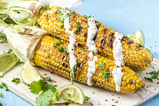 Grilled corn topped with cilantro, lime, and chili flakes on a wooden board.