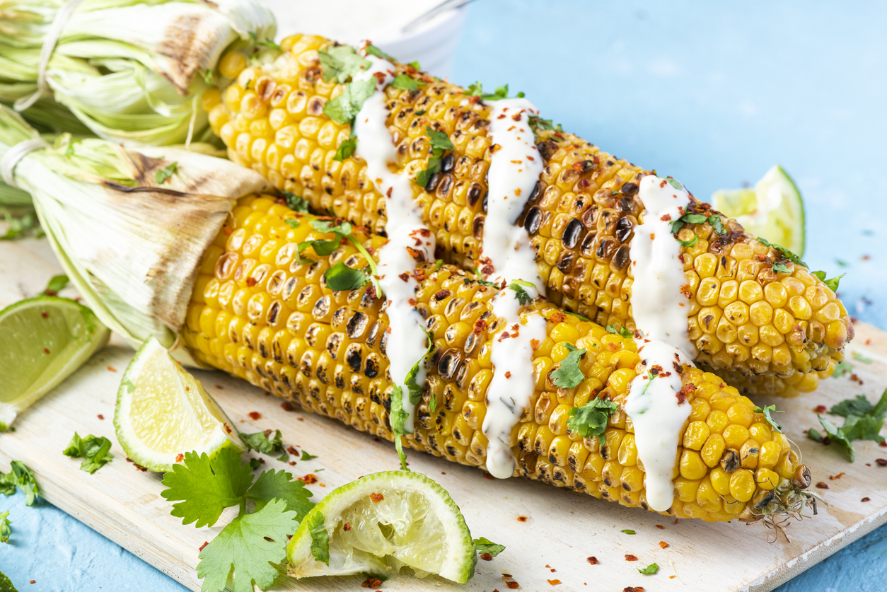 Grilled corn topped with cilantro, lime, and chili flakes on a wooden board.