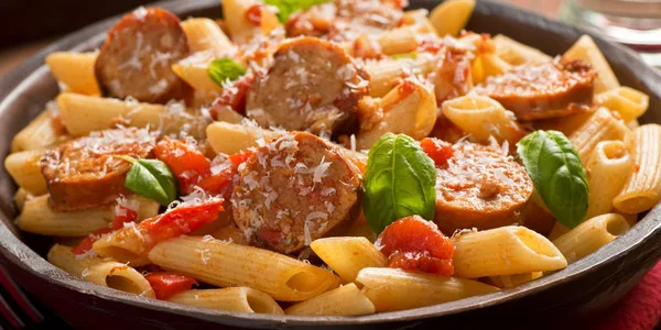 Plated penne pasta dish with sliced Italian sausage, diced tomatoes, fresh basil, and grated cheese for foodservice wholesale distribution.